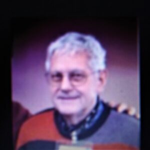 Elderly man with gray hair and glasses wearing a striped sweater.