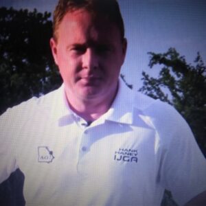 Man in white polo shirt standing outdoors with trees behind him.