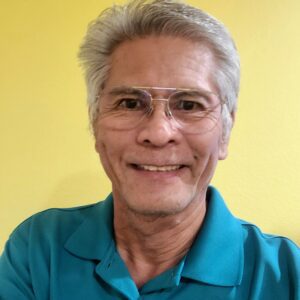 Smiling elderly man in glasses and teal shirt against a yellow background.