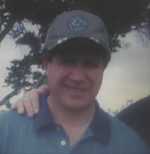 A man wearing a blue cap and shirt outdoors with trees in the background.