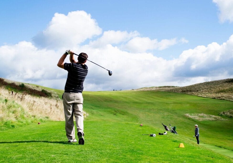 Golfer swinging on a sunny green golf course under a blue sky.